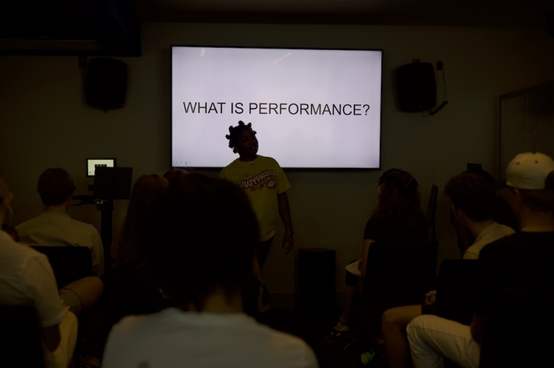 A Tisch Summer High School Recorded Music instructor stands in a dark classroom, backlit, in front of a screen that reads WHAT IS PERFORMANCE? in all-caps